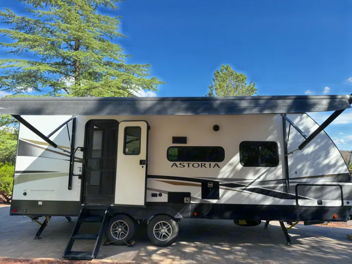 A white & black Astoria travel trailer RV rental, with awning extended, parked at a sunny campsite under a clear blue sky.. Astoria Astoria Trailer 2022
