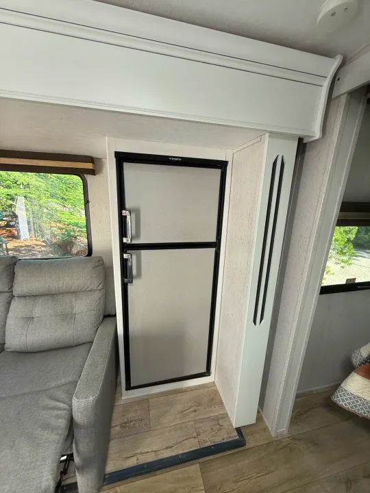 Interior of an RV rental featuring a Dometic refrigerator, a comfortable gray sofa, and a window with a green outdoor view.. Astoria Astoria Trailer 2022