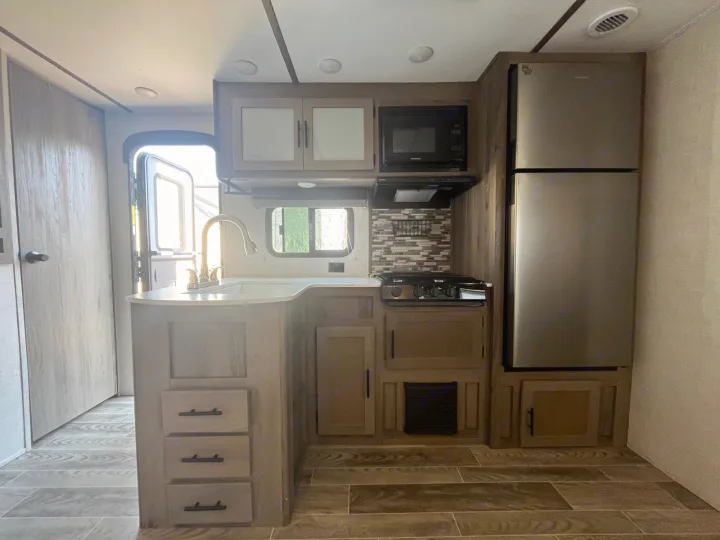Bright RV rental kitchen with wood-look cabinets, stainless fridge, stove, microwave, and sink. Ready for road trip cooking.. Gulf Stream Kingsport 2023
