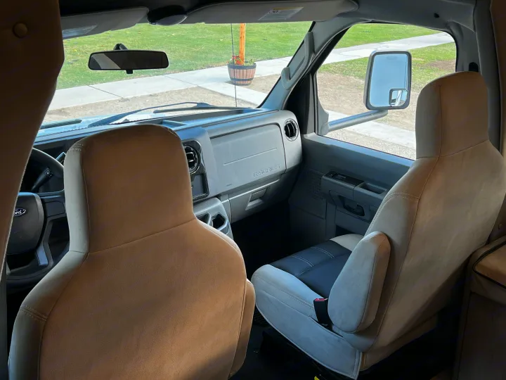 Inside an RV rental's front cabin, showing tan & grey seats, dashboard, and the view through the windshield.. Forest River Coachmen Freelander Premier 2021