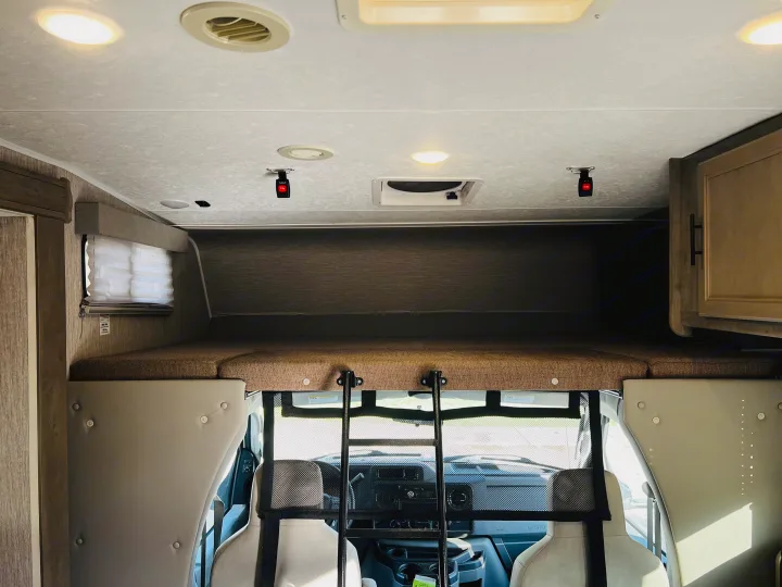 Inside an RV rental: view of the overhead bunk with ladder leading to the front cab. Ceiling lights, vent, and window are visible.. Forest River Coachmen Freelander Premier 2021