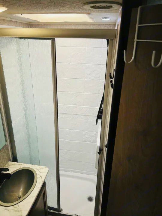 RV rental bathroom with a modern sink, textured glass shower, white tiled interior, overhead lights, and handy towel hooks.. Forest River Coachmen Freelander Premier 2021