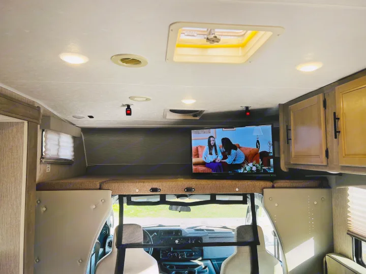 Inside an rv rental: TV playing on an overhead bunk, bright lights, ceiling vent, skylight, and cockpit view with mesh partition.. Forest River Coachmen Freelander Premier 2021