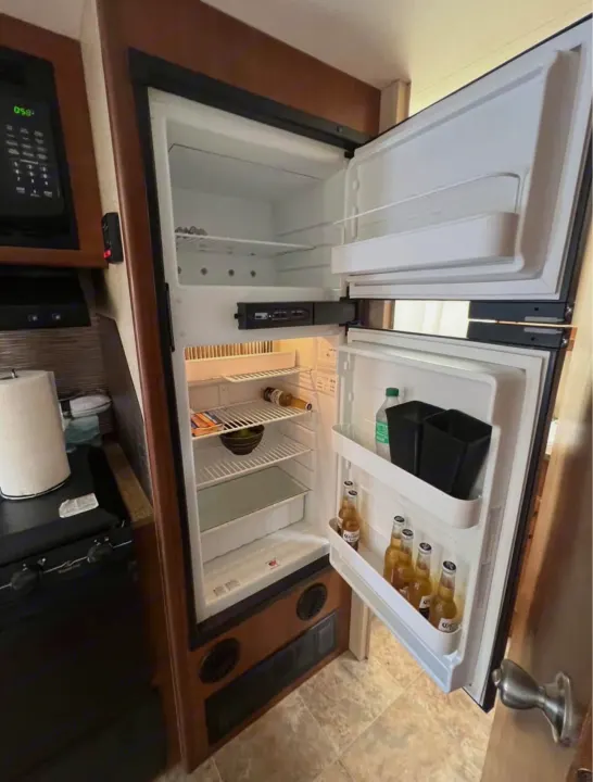 Inside an RV rental: open refrigerator filled with various drinks & items. Microwave, stovetop, and sink are also visible.. Lance Manufacturing Lance Manufacturing Trailer 2016