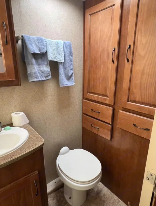 Compact RV rental bathroom with a toilet, sink, and wooden cabinets. Blue towels hang near the vanity.. Lance Manufacturing Lance Manufacturing Trailer 2016