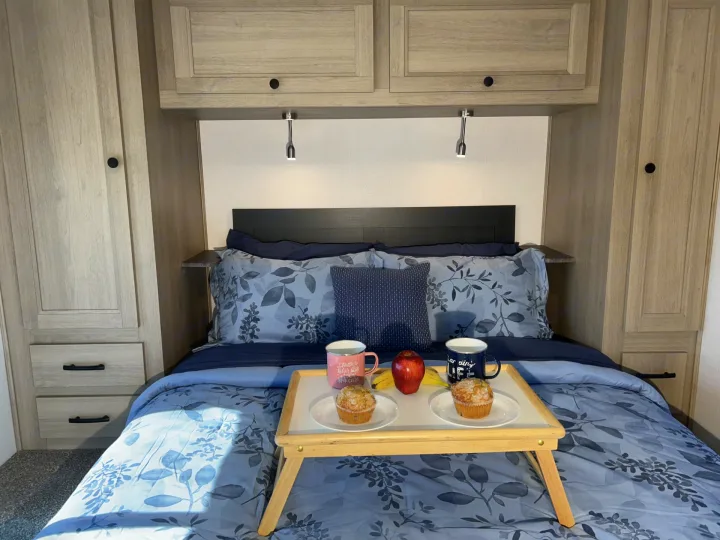 RV rental bedroom with breakfast in bed: muffins, fruit, and mugs on blue floral bedding, plus compact storage.. Forester LE 2026
