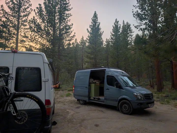 Van life camping in a forest: Two RV rental vans, one with a bike, parked for an outdoor adventure at dusk.. Mercedes-Benz Sprinter 2014