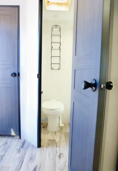 Inside an RV rental bathroom: a white toilet, wall-mounted storage rack, and wood-look floor, viewed through an open doorway.. Venture Venture Trailer 2023