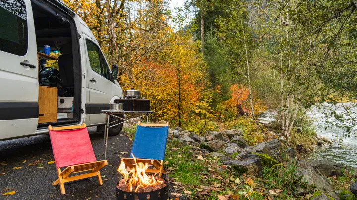 Fall RV rental camping: A camper van beside a vibrant autumn river, with a crackling fire and cozy chairs.. Mercedes Sprinter 2017