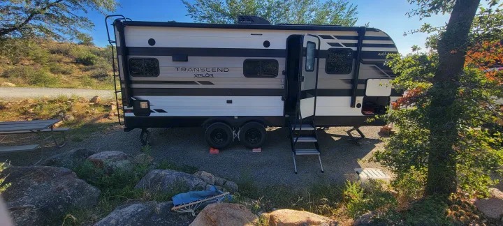 An RV rental: a striped Transcend XPLOR travel trailer, parked at a rocky campsite with open door and steps for easy access.. Grand Design Transcend Xplor 2022