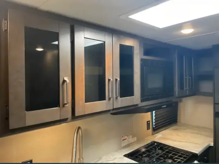 Modern RV rental kitchen: Dark cabinets, glass inserts, built-in microwave, gas cooktop, and sink area.. Grand Design Transcend Xplor 2022