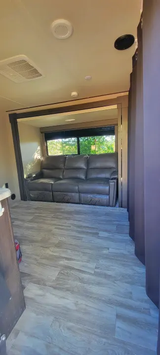 Inside an RV rental: a comfy grey sofa in a slide-out, wood-look floor, and ceiling vents. A welcoming living space.. Grand Design Transcend Xplor 2022