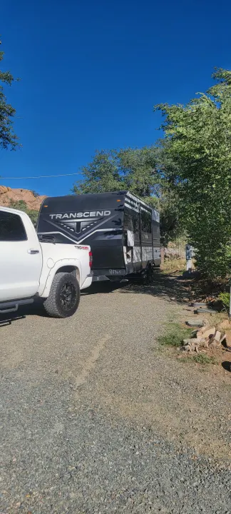 A white pickup truck hitched to a black "Transcend" RV rental on a sunny gravel path, surrounded by trees.. Grand Design Transcend Xplor 2022
