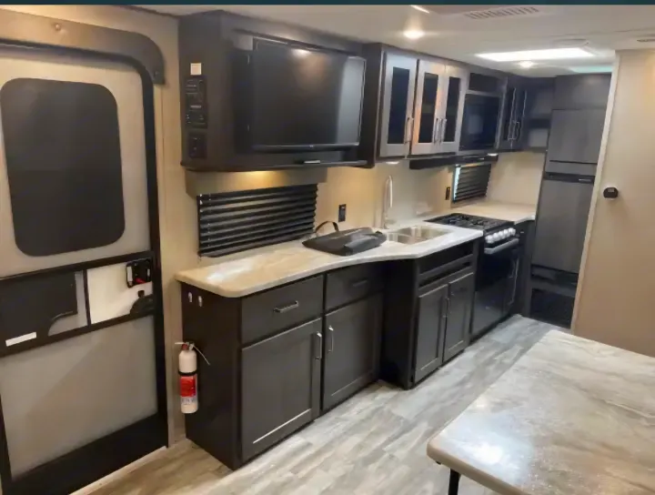 Modern rv rental kitchen featuring dark cabinets, light countertops, sink, stove, fridge, and TV. Entry door and fire extinguisher nearby.. Grand Design Transcend Xplor 2022