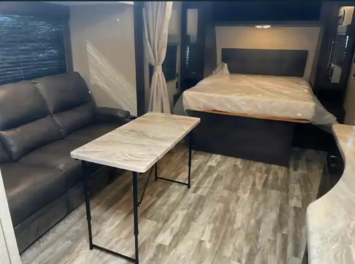 RV rental interior: a dark sofa, light folding table, and a bed with a covered mattress in a modern, compact space.. Grand Design Transcend Xplor 2022