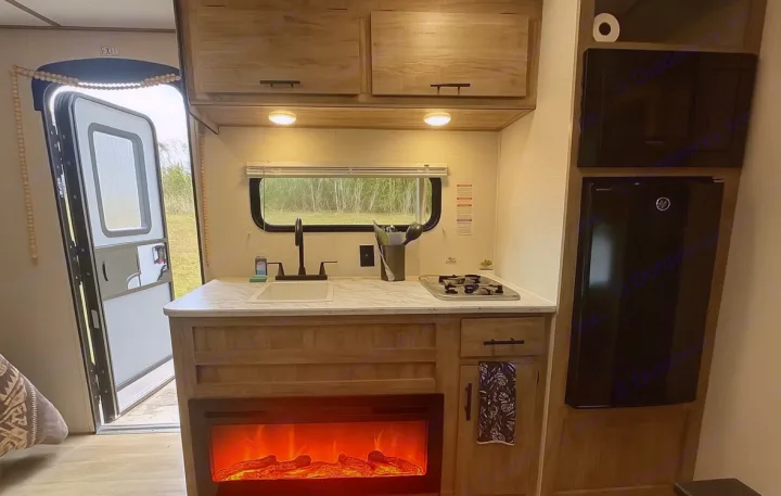 RV rental interior: cozy kitchen with sink, stove, mini-fridge, and an electric fireplace. Open door reveals a natural outdoor scene.. Gulf Stream Enlighten Super Lite 2025