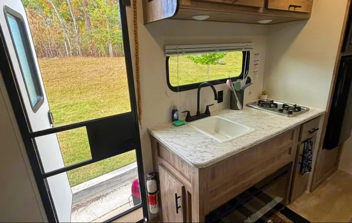 RV rental kitchen with a sink, stovetop, and storage cabinets. An open door reveals a natural outdoor view. Fire extinguisher included.. Gulf Stream Enlighten Super Lite 2025