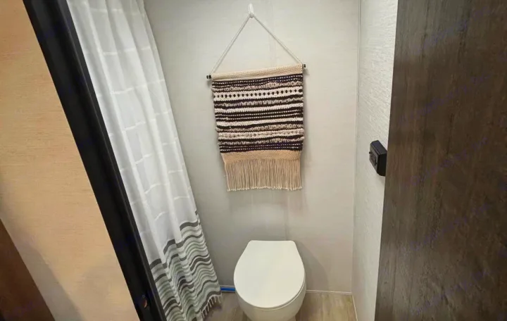This RV rental's compact bathroom features a white toilet, a striped shower curtain, and a decorative tapestry above.. Gulf Stream Enlighten Super Lite 2025