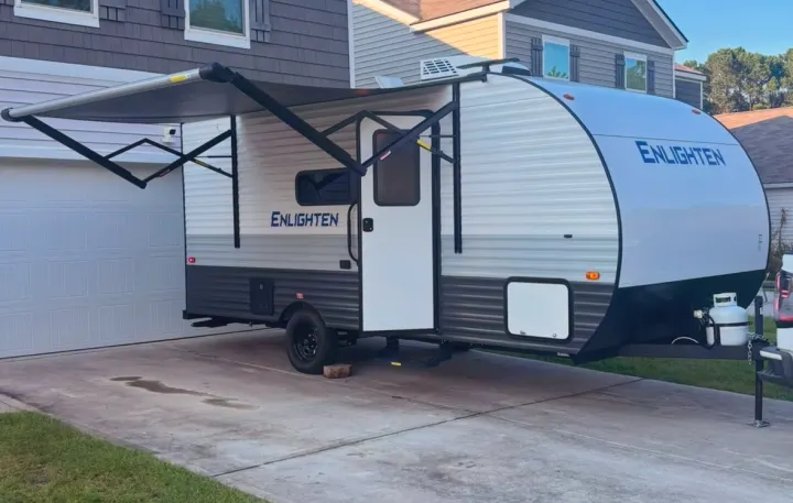 A white & gray 'ENLIGHTEN' travel trailer with an extended awning, parked on a driveway, ideal for an RV rental getaway.. Gulf Stream Enlighten Super Lite 2025