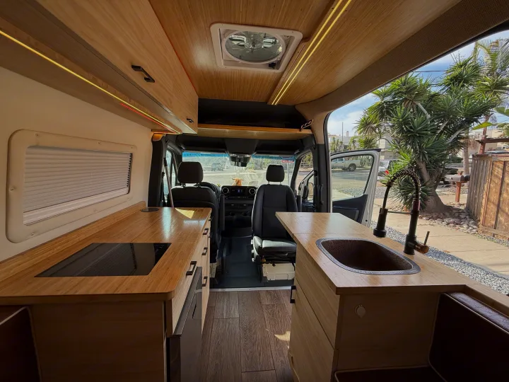 This RV rental's inviting interior features light wood kitchen counters, a copper sink, cooktop, and overhead cabinets with strip lighting.. Mercedes-Benz Sprinter 2024
