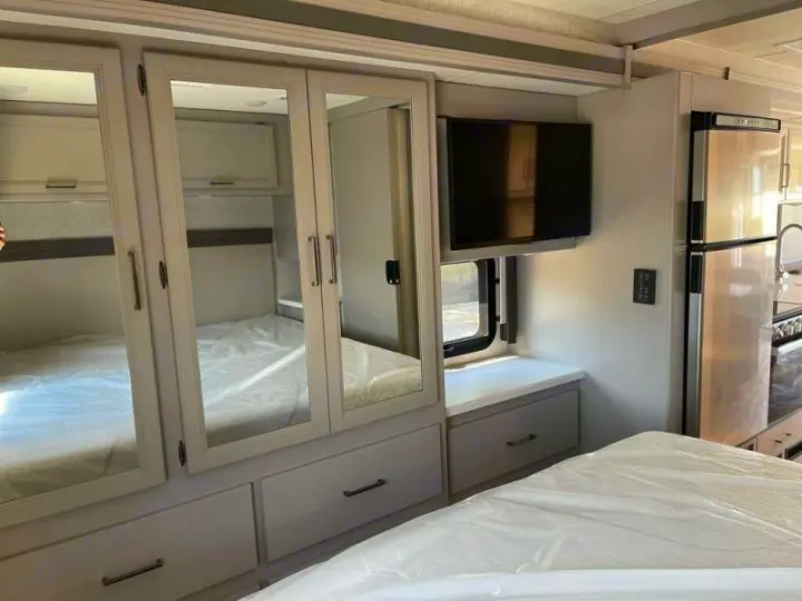 Explore this modern RV rental's bedroom with mirrored wardrobes, TV, and adjacent kitchen area for comfortable travel.. Thor Windsport 2023