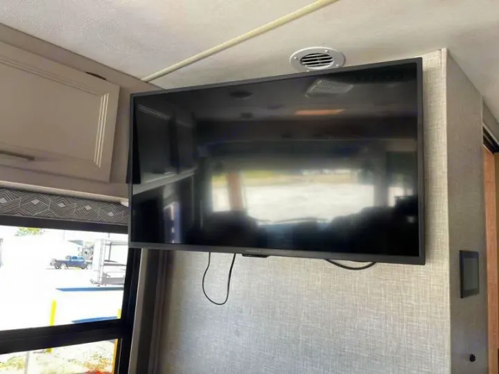 RV rental interior featuring a mounted flat-screen TV, overhead cabinet, and window overlooking a parking lot.. Thor Windsport 2023
