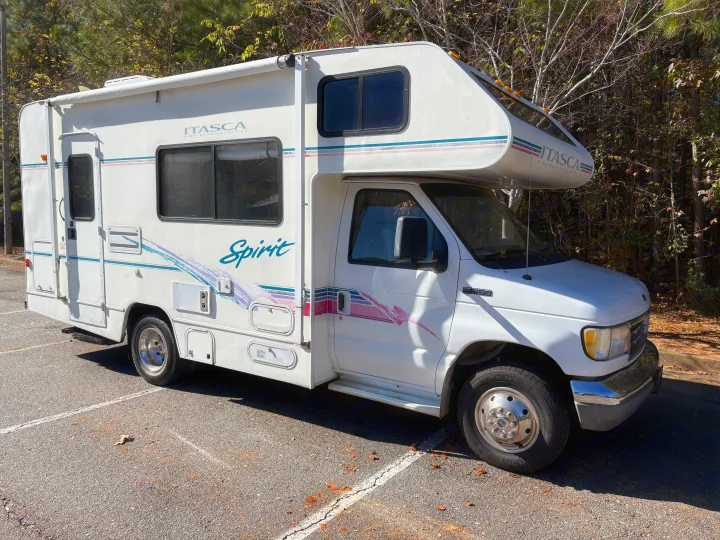 A white Itasca Spirit Class C RV rental is parked on asphalt with trees in the background, showcasing its classic design.. Itasca Spirit 1996
