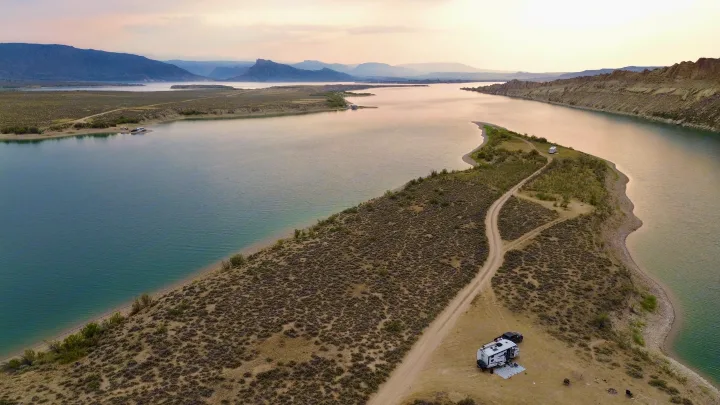 Aerial view of an RV rental campsite on a remote lake peninsula. A white RV & truck are parked by calm waters & distant mountains.. Crossroads RV Sunset Trail Super Lite 2021