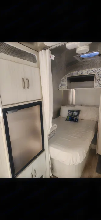 Bright rv rental interior features a cozy bed with white linens, a compact fridge, and light wood-grain cabinets.. Airstream Bambi 2023