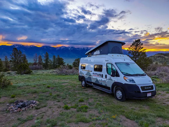 An RV rental camper van with a pop-top enjoys a stunning mountain sunset vista from a grassy campsite with a fire pit.. Winnebago Solis 2022