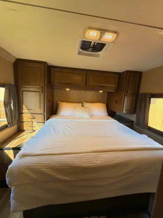 Cozy RV rental bedroom featuring a comfy bed with white linens, overhead cabinets, a ceiling light, and side windows.. Thor Majestic 2019