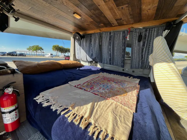 Cozy RV rental interior with a comfortable bed, fan, and scenic view. A fire extinguisher is visible for safety.. Chevrolet Astro 1992