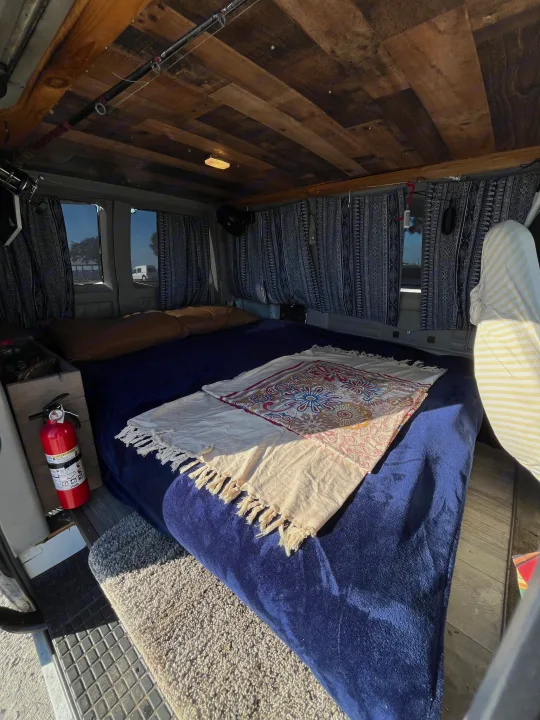 Inside a cozy RV rental: a plush bed, wooden ceiling, fishing rods, and patterned curtains create a warm van conversion.. Chevrolet Astro 1992