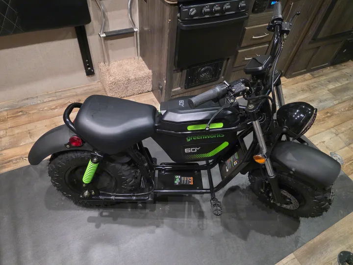 A black Greenworks 60V electric mini-bike with green accents, parked inside an rv rental, showcasing its portable design.. Keystone Coleman 2025