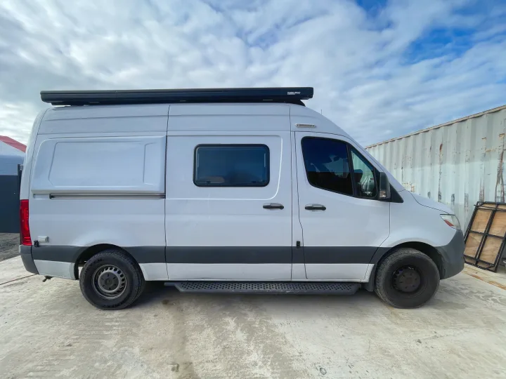 This white RV rental camper van features a rooftop awning, side step, and tinted windows, parked on concrete under a cloudy sky.. MERCEDES SPRINTER 144 2020