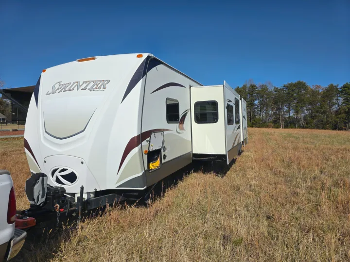 This Sprinter RV rental travel trailer with extended slide-outs rests in a sunlit, dry field under a clear blue sky.. Keystone RV Sprinter 2013
