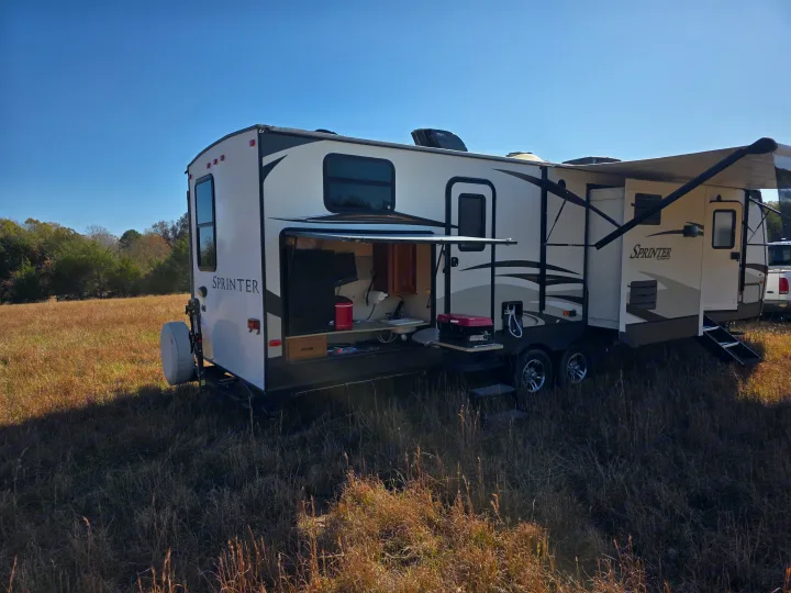 This Sprinter rv rental travel trailer, with its open outdoor kitchen, is perfectly set up for camping in a sunny, dry grassy field.. Keystone RV Sprinter 2013
