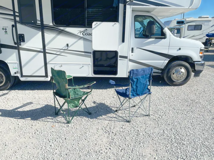 Enjoying the outdoors with an RV rental! A white Class C motorhome with open storage and two chairs on a gravel site.. Forest River Coachmen Leprechaun 2025