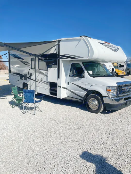 White Class C rv rental parked on gravel with awning extended, two camping chairs nearby. Ready for your next adventure!. Forest River Coachmen Leprechaun 2025