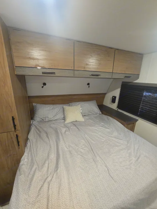RV rental bedroom with light wood overhead storage, a comfortable striped bed, pillows, and bedside lighting.. Forest River Campsite Reserve 2025