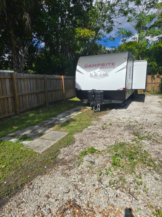A white Campsite Reserve RV rental with its door open, parked on a gravel lot beside a wooden fence and green trees.. Forest River Campsite Reserve 2025