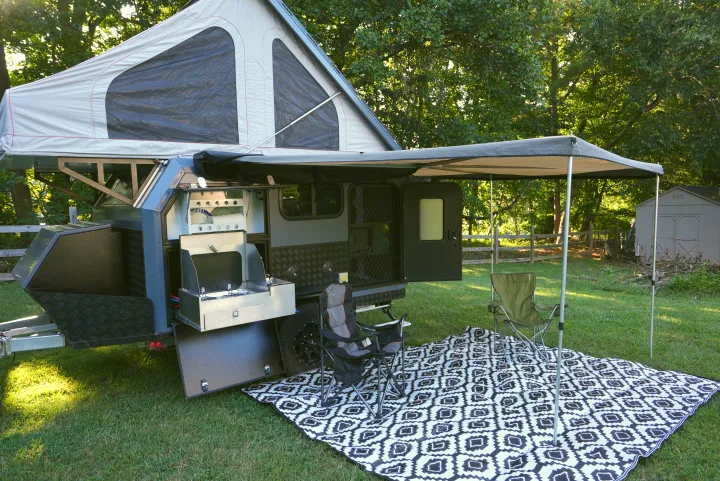 This grey and black rv rental pop-up camper is set up outdoors with an extended awning, outdoor kitchen, chairs, and mat.. Zoomies Campers Spirit 2025