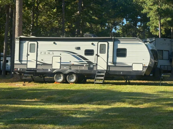 A grey & white Puma travel trailer rv rental, set up on grass under pine trees at a sunny campground.. Forest River Palomino Puma 2022