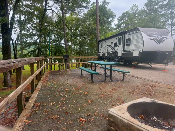 RV rental campsite with a Palomino Puma travel trailer, green picnic table, fire pit, and trees, offering a distant water view.. Forest River Palomino Puma 2022