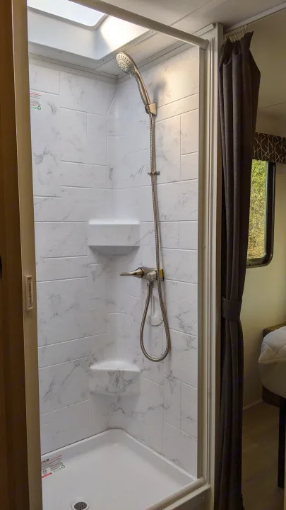 Clean, compact shower in an RV rental, featuring white marble-look walls, handheld showerhead, and a glimpse of nature outside.. Forest River Sunseeker 2025
