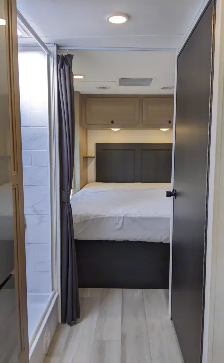 View of an RV rental interior, featuring a cozy bedroom with white bedding, dark headboard, and an adjacent shower stall.. Forest River Sunseeker 2025