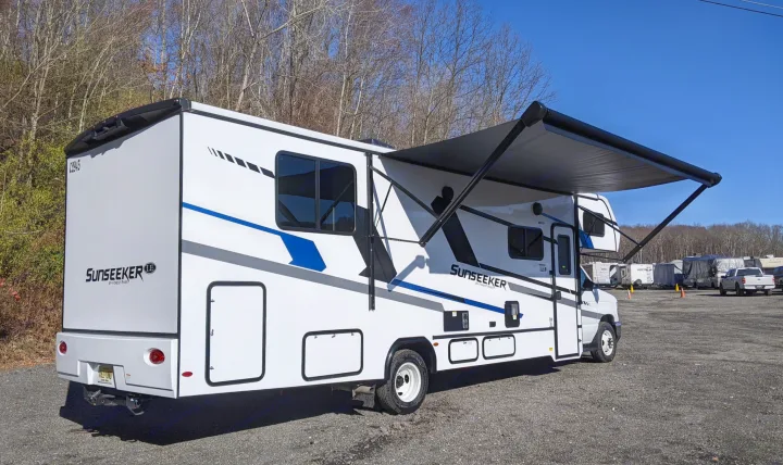 A white Sunseeker LE motorhome RV rental with blue/gray graphics and an extended dark awning on a gravel lot.. Forest River Sunseeker 2025