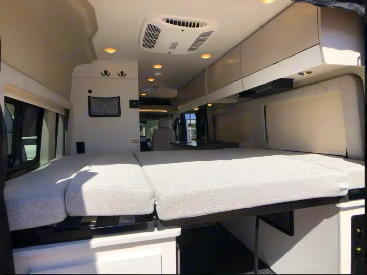 Cozy RV rental interior with a bed featuring chevron cushions, overhead cabinets, ceiling lights, and an AC unit.. Entegra Coach Ethos 2025