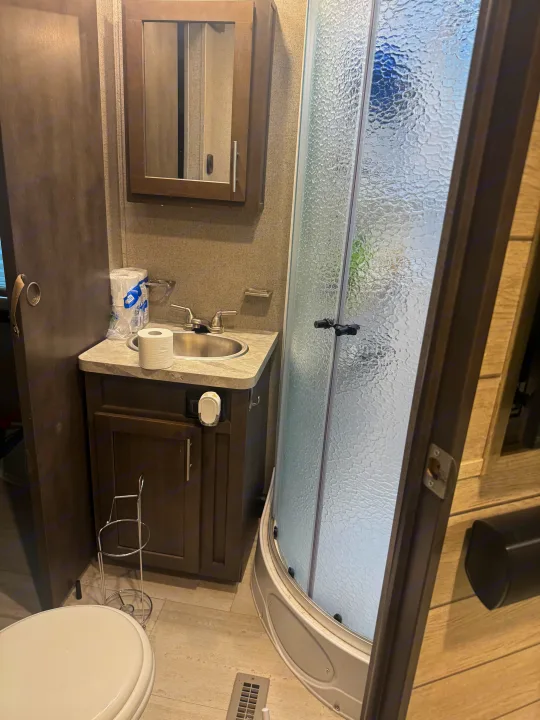 This compact RV rental bathroom features a sink with a mirrored cabinet above, a toilet, and a textured glass shower stall.. Forest River Flagstaff Classic 2020