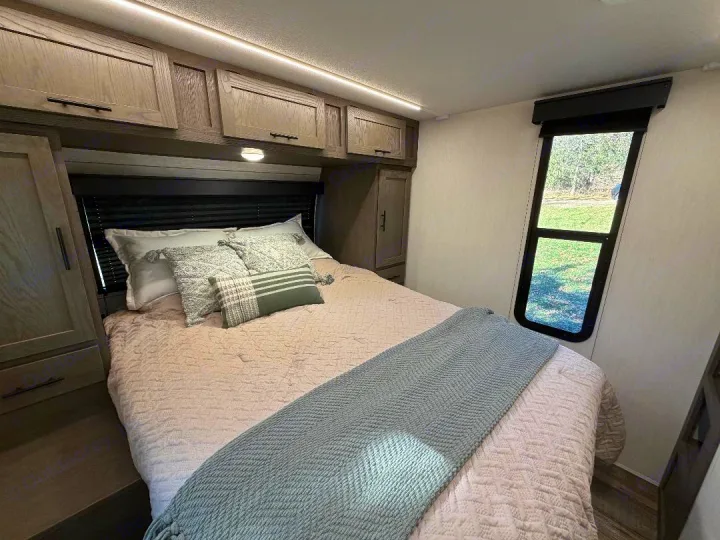 Cozy RV rental bedroom with overhead storage, a comfy bed, and a window view overlooking green scenery.. Forest River Cherokee Alpha Wolf 2025